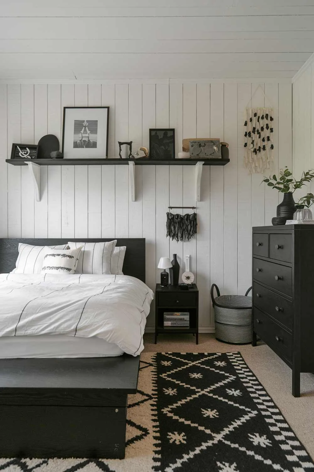 A cozy black japandi bedroom interior design. The room has a black wooden platform bed with white bedding. There's a black dresser and a shelf with decorative items. The walls have white painted wood panels. The floor is covered with a black and white patterned rug.