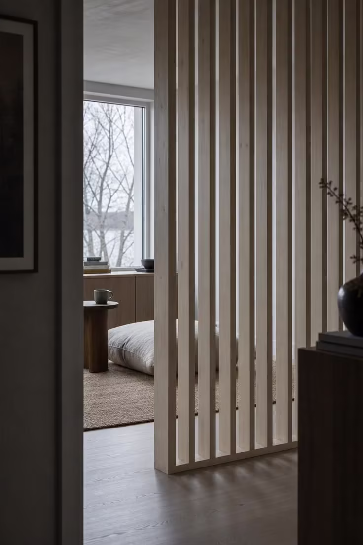 13 Gentle Japandi Reading Nook Ideas for Restful Rooms 5 A birch slat room divider framing a low walnut stool and floor cushion in a cool-lit japandi reading nook viewed from a doorway perspective.