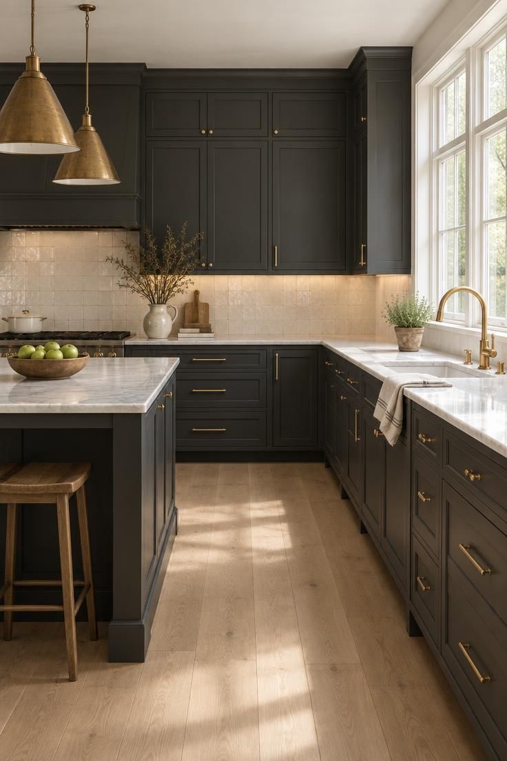 A wide view of a completed charcoal kitchen with pale marble counters, warm white tile, oak floors, and brass pendants over a dark island.