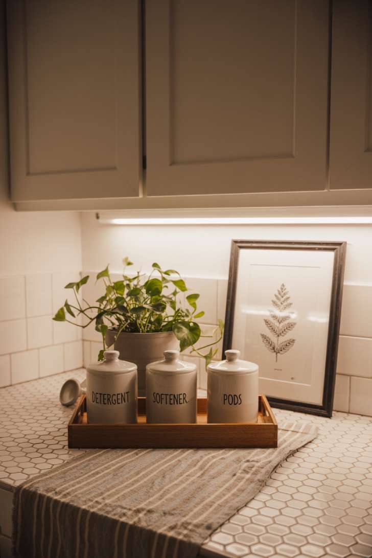 13 Simple Laundry Room Decor Ideas for a Beautiful Space 10 A styled laundry room countertop with ceramic labeled jars, a trailing pothos, and a framed botanical print on white shaker cabinetry