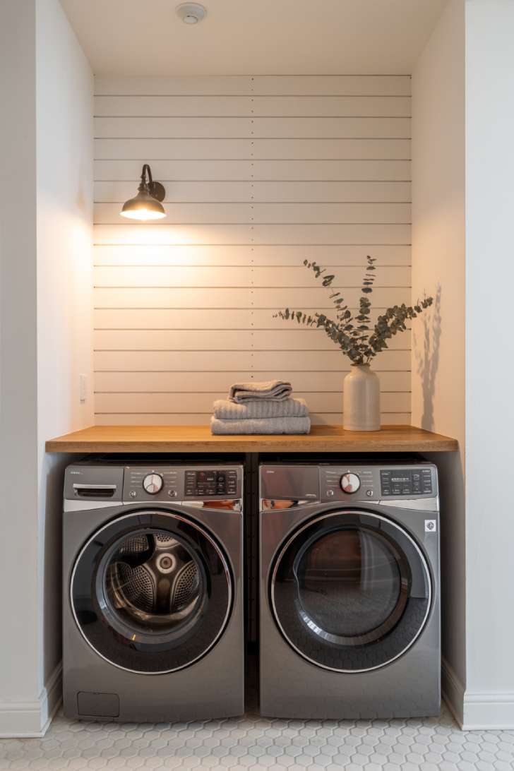 11 Beautiful Laundry Room Wall Decor Ideas That Inspire 3 A white shiplap accent wall behind a washer and dryer, with a black wall sconce, wooden countertop, folded towels, and dried eucalyptus in a white vase.