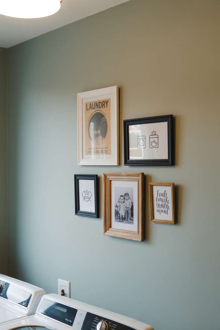 11 Beautiful Laundry Room Wall Decor Ideas That Inspire 5 A gallery wall of five mixed frames on a soft gray wall above a washer, featuring vintage prints, laundry symbols, a family photo, and a hand-lettered quote.