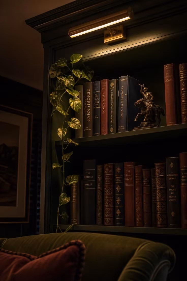 What to Check Before Styling a Living Room With Bookshelves for the First Time 4 A close-up of a dark green built-in bookshelf at night with a brass picture light casting warm light across book spines and a bronze sculpture.