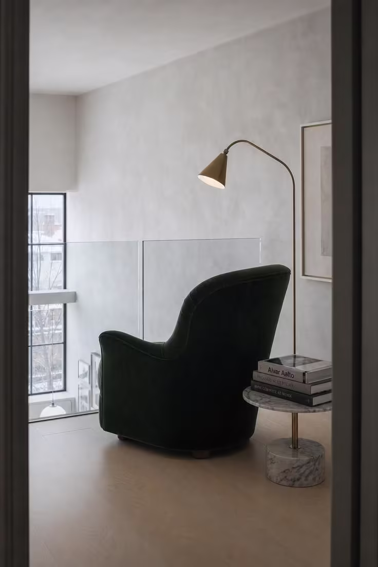 11 Inviting Loft Reading Nook Ideas for Every Home 7 A loft nook with a frameless glass panel railing, forest green velvet chair, brass arc lamp, and cool overcast light from below.