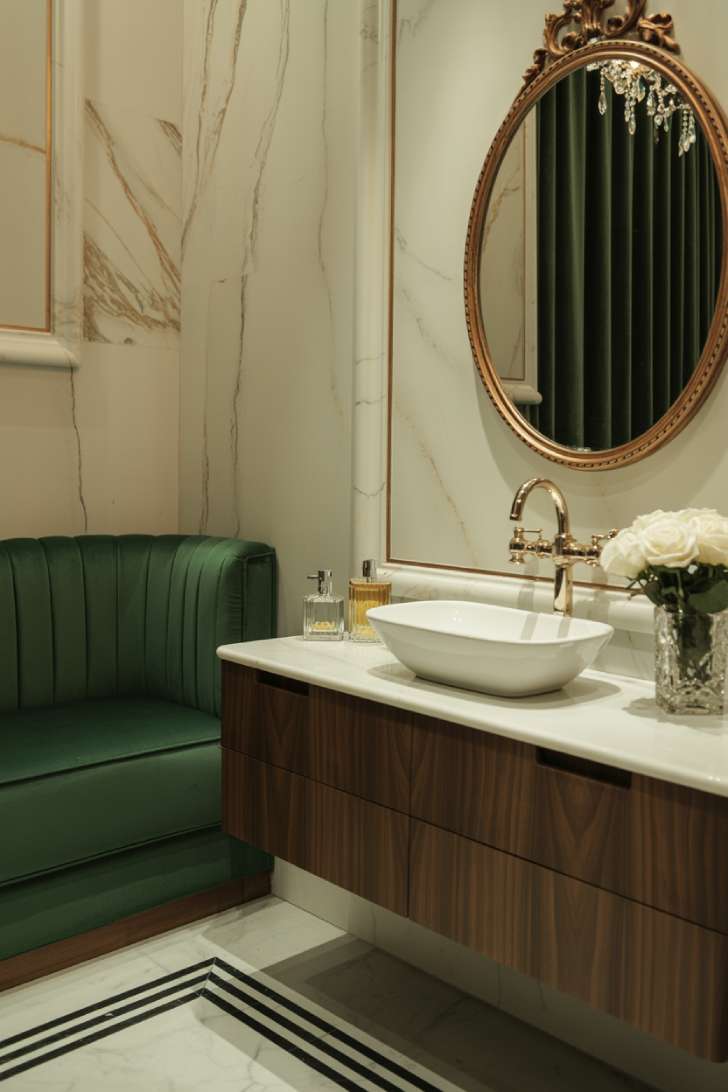 Luxury powder room with cream and gold marble walls, dark walnut floating vanity, white vessel sink, brass fixtures, oval gold mirror, crystal chandelier, emerald velvet seating, and geometric marble flooring
