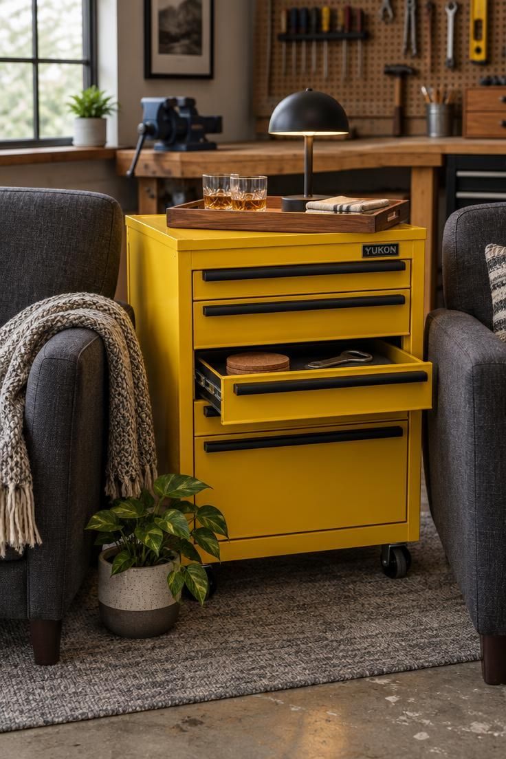 13 Cozy Man Cave Garage Workshop Ideas for Hangout & Build 4 Close-up of a bright yellow rolling tool cabinet used as a side table between two charcoal club chairs, topped with a tray of glasses and a small lamp, with a workbench softly blurred behind.