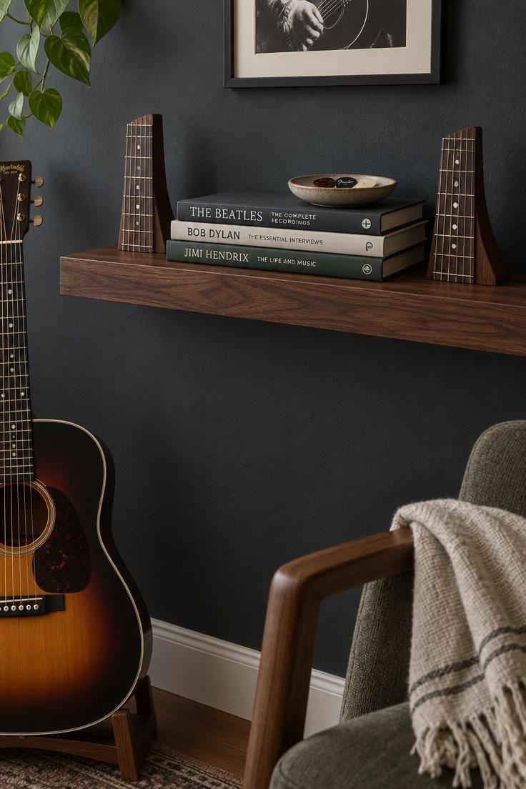A close-up of a walnut floating shelf with fretboard-shaped bookends, a stack of music books, a ceramic pick dish, and the neck of a sunburst acoustic guitar entering from the lower left.