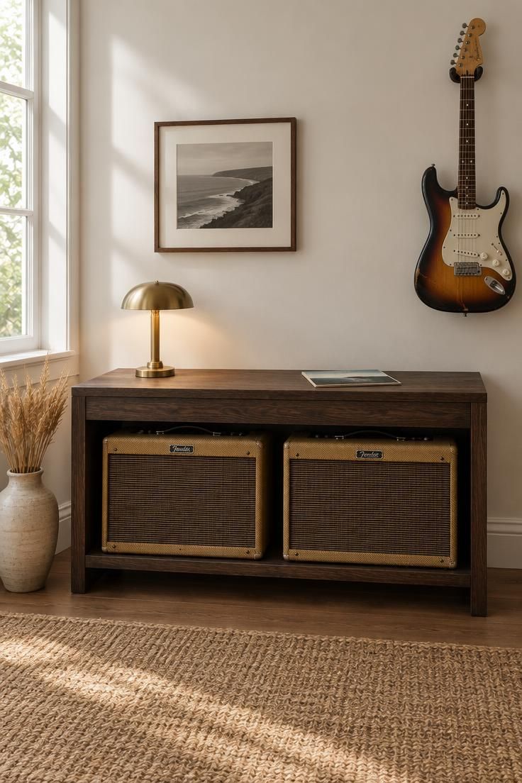 A low dark oak console with two tweed amplifiers on its lower shelf, a brass lamp and vinyl record sleeve on top, and an electric guitar hanging on the wall to the right.