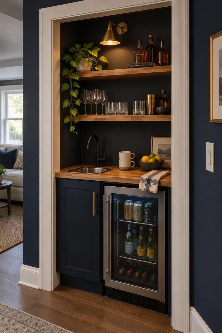 9 Warm Man Cave Kitchen Ideas for Easy Game-Day Hosting 3 A converted closet micro-kitchen with warm white interior paint, a narrow butcher block counter, a small stainless bar sink with matte black faucet, open shelves of glassware and amber bottles, and a brass wall lamp, set in a deep navy wall.