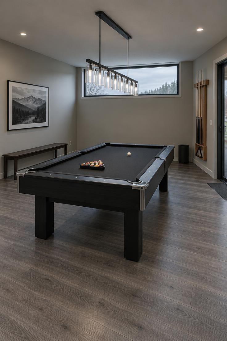 Modern residential billiard room with a black slate pool table and black rails, a long black iron linear pendant with glass tubes over it, pale gray walls, a natural wood wall-mounted cue rack, a slim dark wood bench along one wall, and clear space on all sides of the table.
