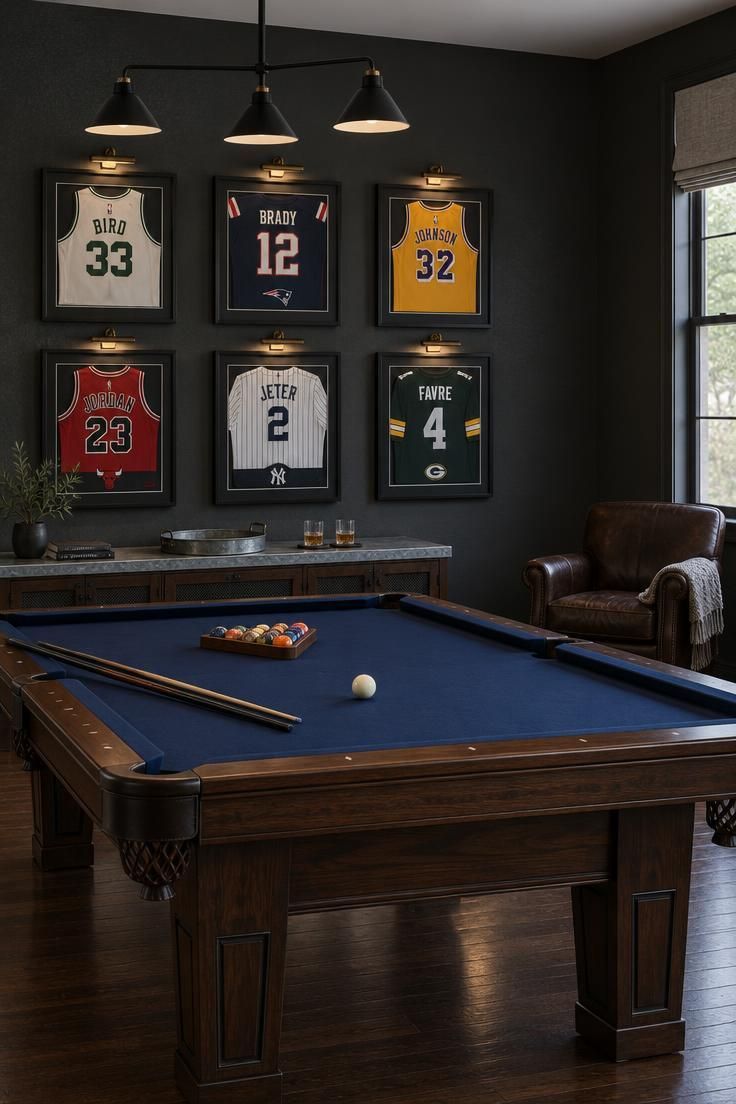 Residential game room with a navy felt pool table and dark walnut rails, six black-framed jerseys in two rows on a charcoal wall each lit by a picture light, a galvanized-tin-topped console below, a dark leather club chair in the corner, and a three-arm black pendant over the table.