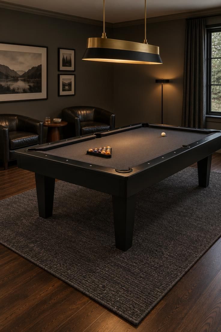 Residential pool room with a charcoal felt table under a brass and black bow-front pendant, two black leather club chairs with a round walnut side table along a side wall, a charcoal wool rug under the table with a visible border, dark wood flooring, and deep warm gray walls.
