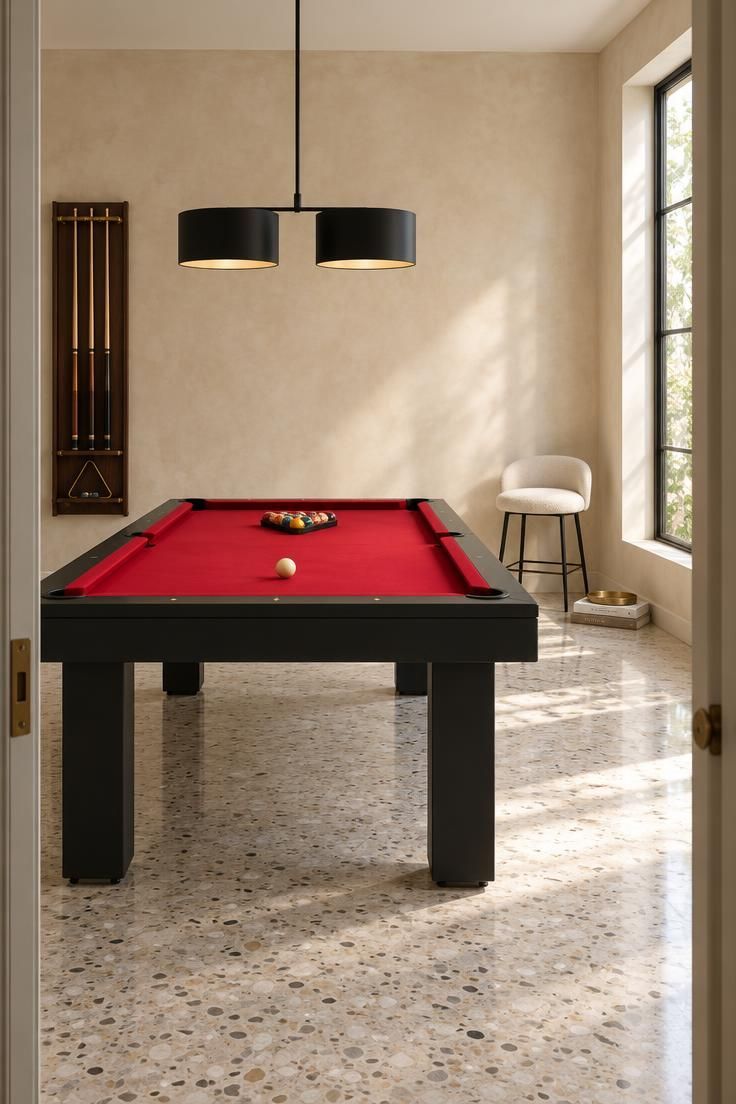 Bright residential billiard room with a red felt pool table and matte black rails on a pale terrazzo-look floor, a cream plaster wall behind, a black double-drum pendant over the table, an off-white boucle stool near the wall, and a dark wood cue rack with brass hardware on the side wall.