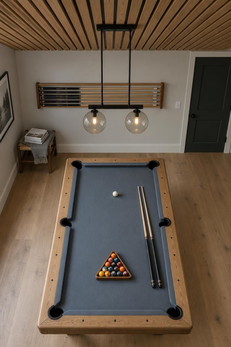 Overhead view of a residential pool room with a blue gray felt table and natural wood rails under a stained birch slat ceiling with a black and glass two-light pendant, a long horizontal wall-mounted cue rack storage line near the entry door, white oak flooring, and a small woven-seat wooden stool in the corner.