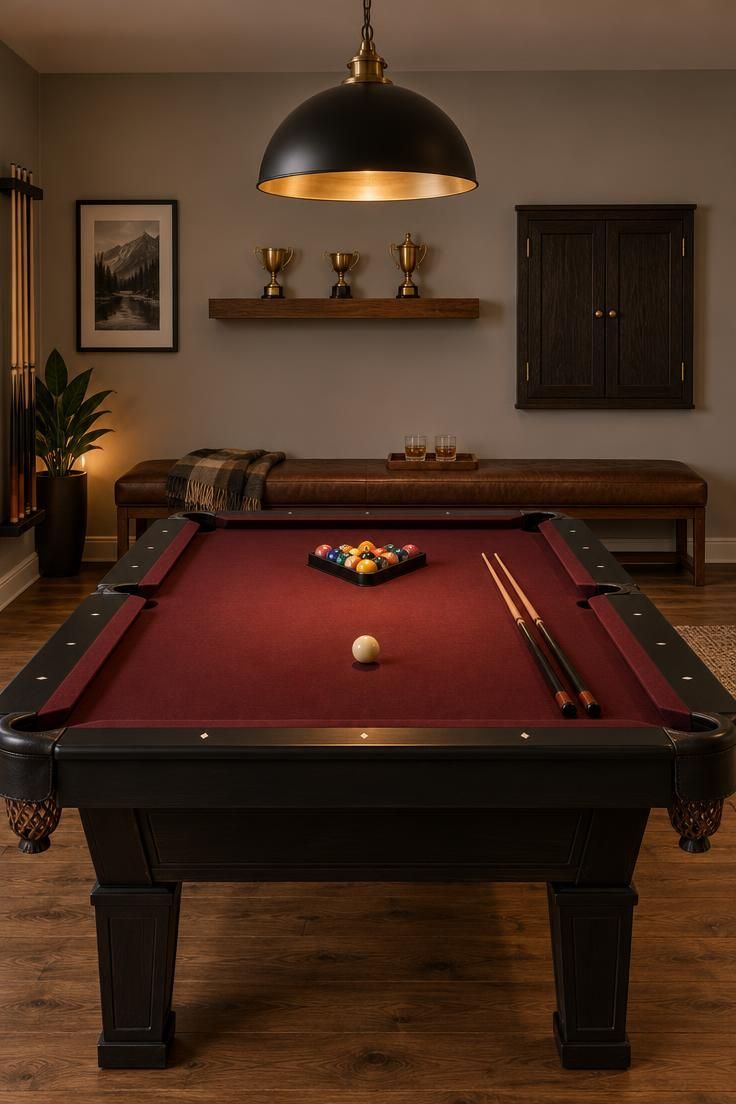 Residential man cave pool room with a burgundy felt table and black rails under a black metal dome pendant with a brass inner finish, a floating walnut shelf with three brass trophies above a closed dark stained dart cabinet, a low leather bench beneath, warm light gray walls, and medium brown wood flooring.