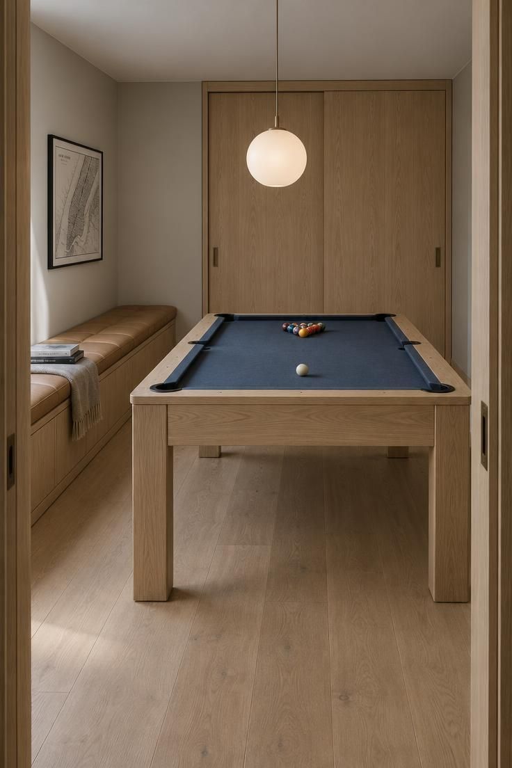 Small residential pool room with a pale oak pool table and dark slate blue felt, a long low bench with a padded tan seat along the side wall, a floor-to-ceiling cabinet with two closed sliding oak doors on the back wall, a single frosted glass globe pendant over the table, pale oak flooring, and a small black and white framed map above the bench.