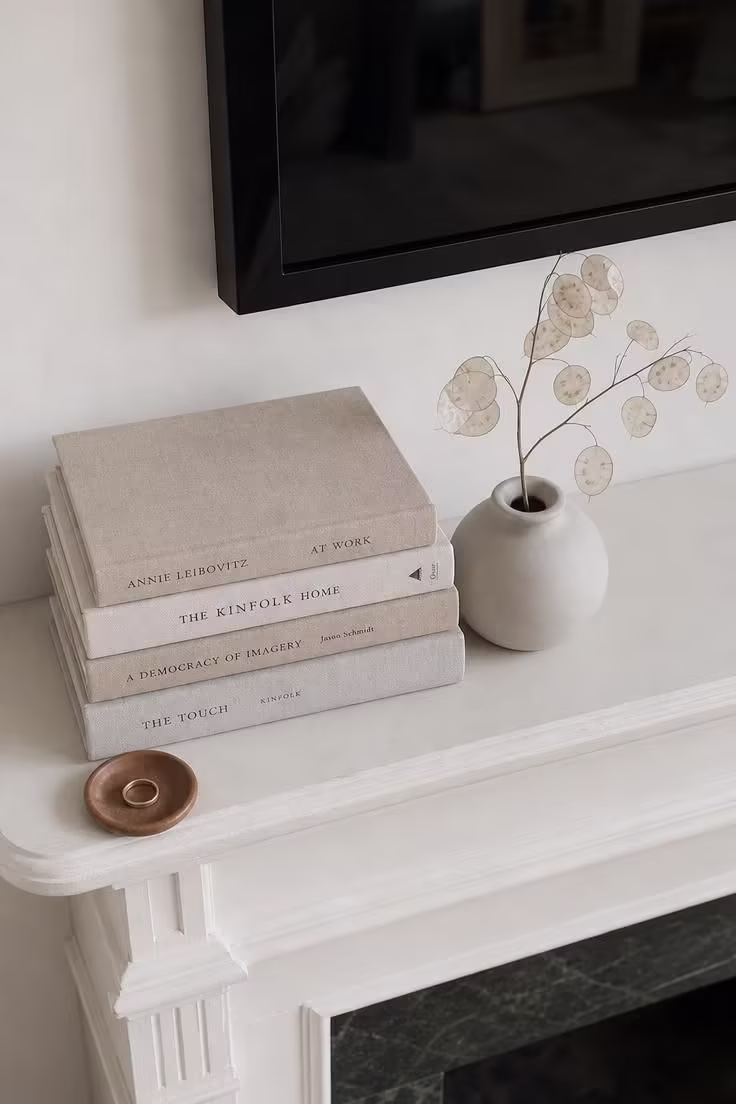 Linen-wrapped hardcover books stacked beside a matte ceramic vase with dried lunaria on a white wood mantel below a TV