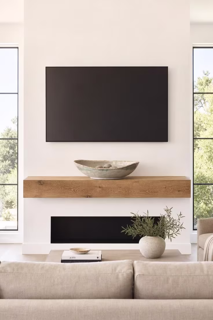 A large handmade stoneware bowl centered on a natural oak mantel shelf beneath a wall-mounted TV in a bright minimalist room