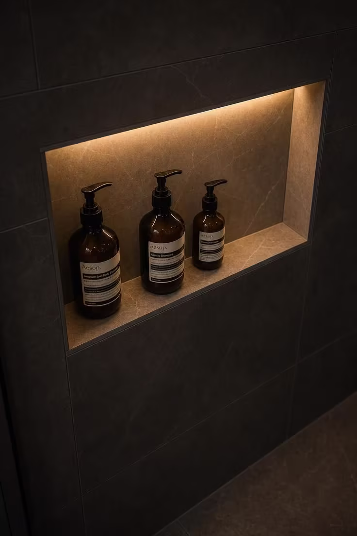 9 Low-Lift Updates That Make Master Bathrooms With Walk In Showers Feel Like a Spa Retreat 3 A recessed shower niche illuminated by a warm LED strip light, casting a soft glow over amber glass bottles against dark charcoal tile
