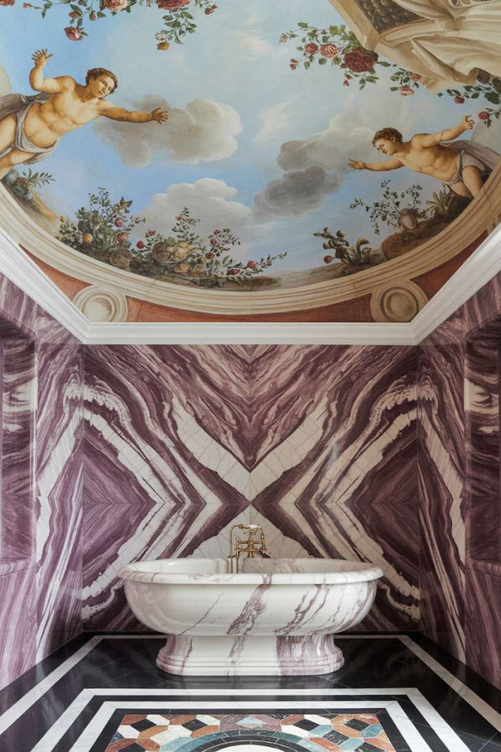 An Italian palazzo–inspired maximalist bathroom with a barrel-vaulted frescoed ceiling, bookmatched Calacatta Viola marble walls, a travertine freestanding tub, and a full-wall antique mirror vanity