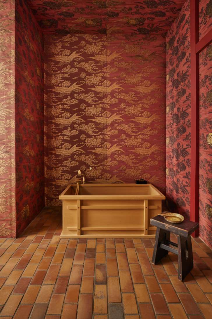 A Far Eastern–inspired maximalist bathroom with deep lacquer red wallpaper, an antique terracotta herringbone floor, a hinoki cypress ofuro tub, and a carved cinnabar lacquer vanity