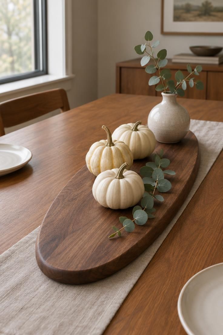 12 Warm Mid-Century Modern Thanksgiving Table Ideas 4 Eye-level view of an oval walnut serving board on a teak table holding three cream gourds, a eucalyptus sprig, and a small rounded vase, with open space around it.