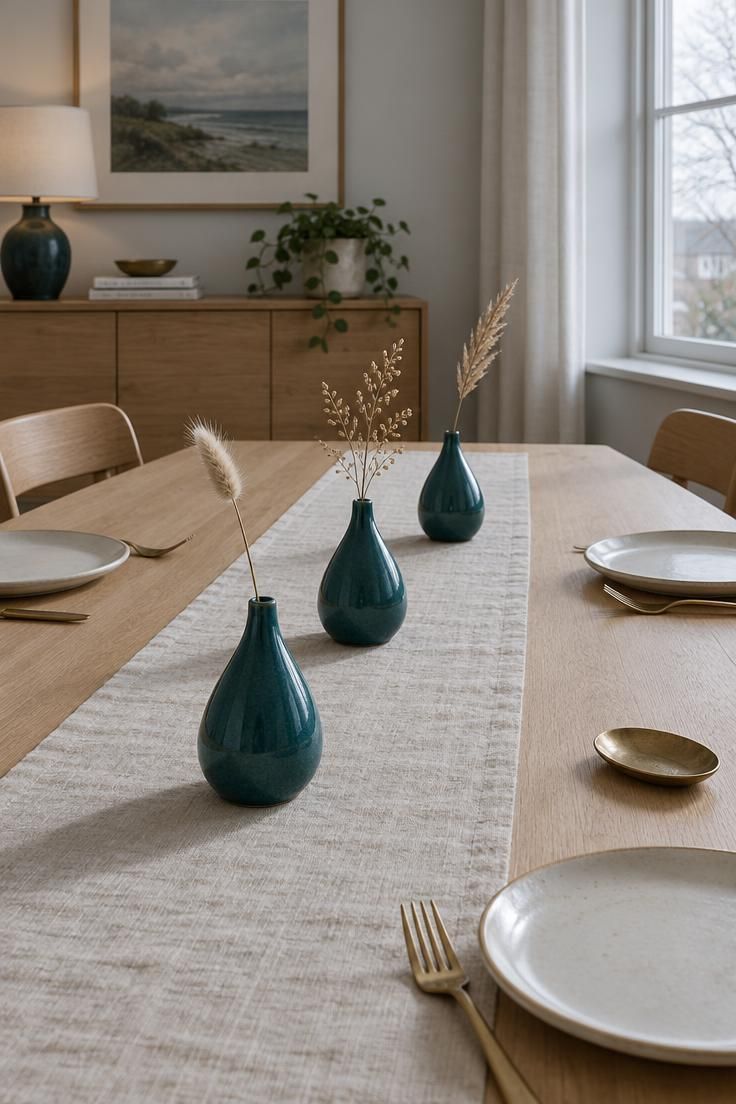 12 Warm Mid-Century Modern Thanksgiving Table Ideas 8 Eye-level view of three deep teal ceramic bud vases with dried grass stems on a light oak table, with cream plates and brass forks.