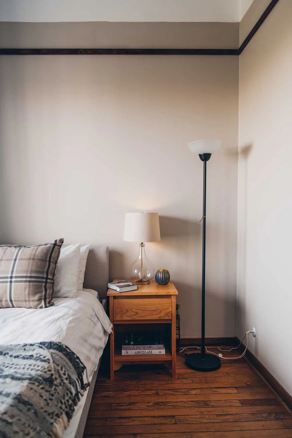 A cozy minimalist apartment bedroom with a queen-size bed, a small nightstand, and a floor lamp. The nightstand has a lamp and a book. The walls are painted beige, and there is a strip of wood along the top of the wall. The floor is made of hardwood.