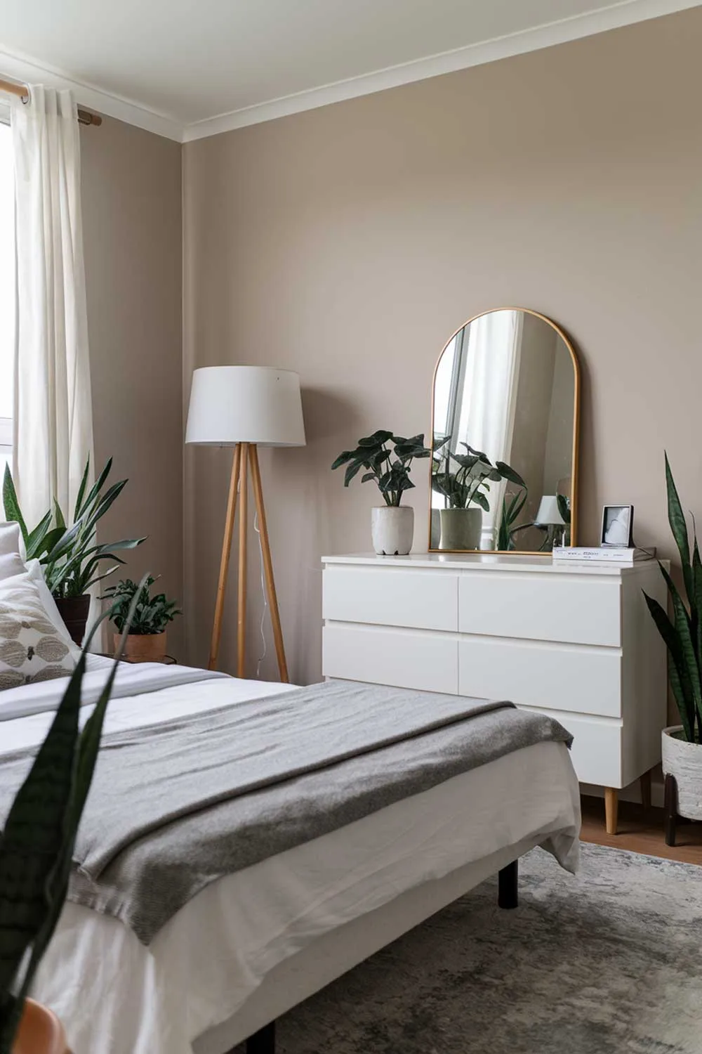 A cozy and beautifully designed minimalist apartment bedroom. The room has a queen bed with a white sheet and a gray blanket. There is a white dresser with a mirror and a few items on top. A white lamp with a gray base stands next to the bed. The walls are painted beige. The floor is covered with a gray rug. The room has a few potted plants.