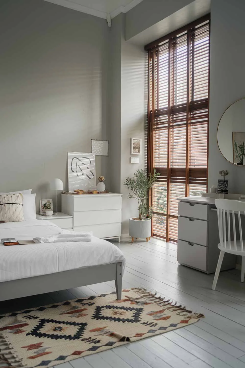 A cozy and beautifully designed minimalist apartment bedroom. The room has a white bed with a gray frame, a white dresser with a gray drawer, and a white chair. There is a small potted plant near the window. The walls and floors are painted in light gray. There is a rug with a geometric pattern near the bed. The room has a large window with wooden blinds.