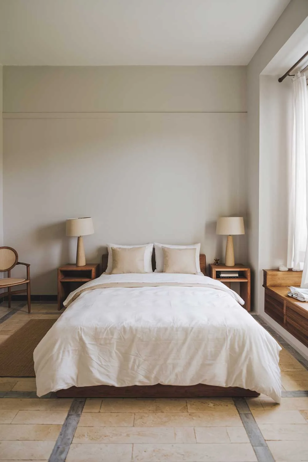 A cozy and beautifully designed minimalist apartment bedroom. The room has a queen-sized bed with a white duvet and two beige pillows. There is a wooden bedside table on either side of the bed, each with a beige lampshade. The walls are painted in a light gray hue. The floor is made of beige and gray tiles. There's a wooden chair in the corner near the window. The window has a white curtain.