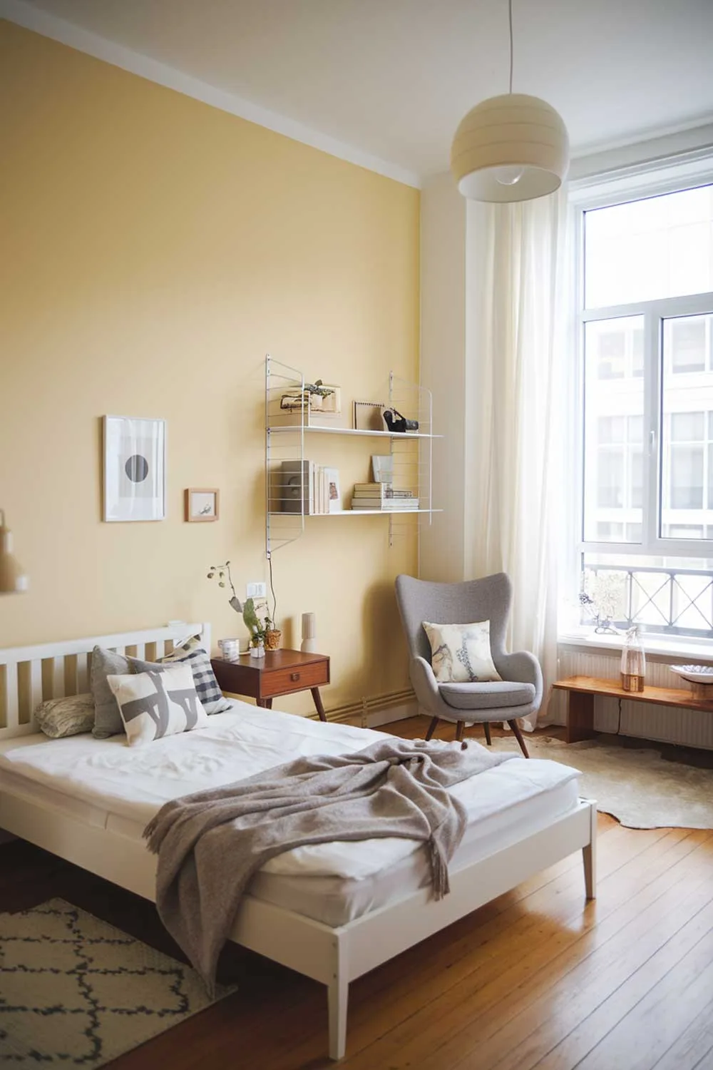 A cozy and beautifully designed minimalist apartment bedroom with a white bed frame, a wooden nightstand, a gray chair, a white wall shelf, and a few decorative items. The room has a soft yellow accent wall and is illuminated by a large window and a hanging lamp. The flooring is hardwood.