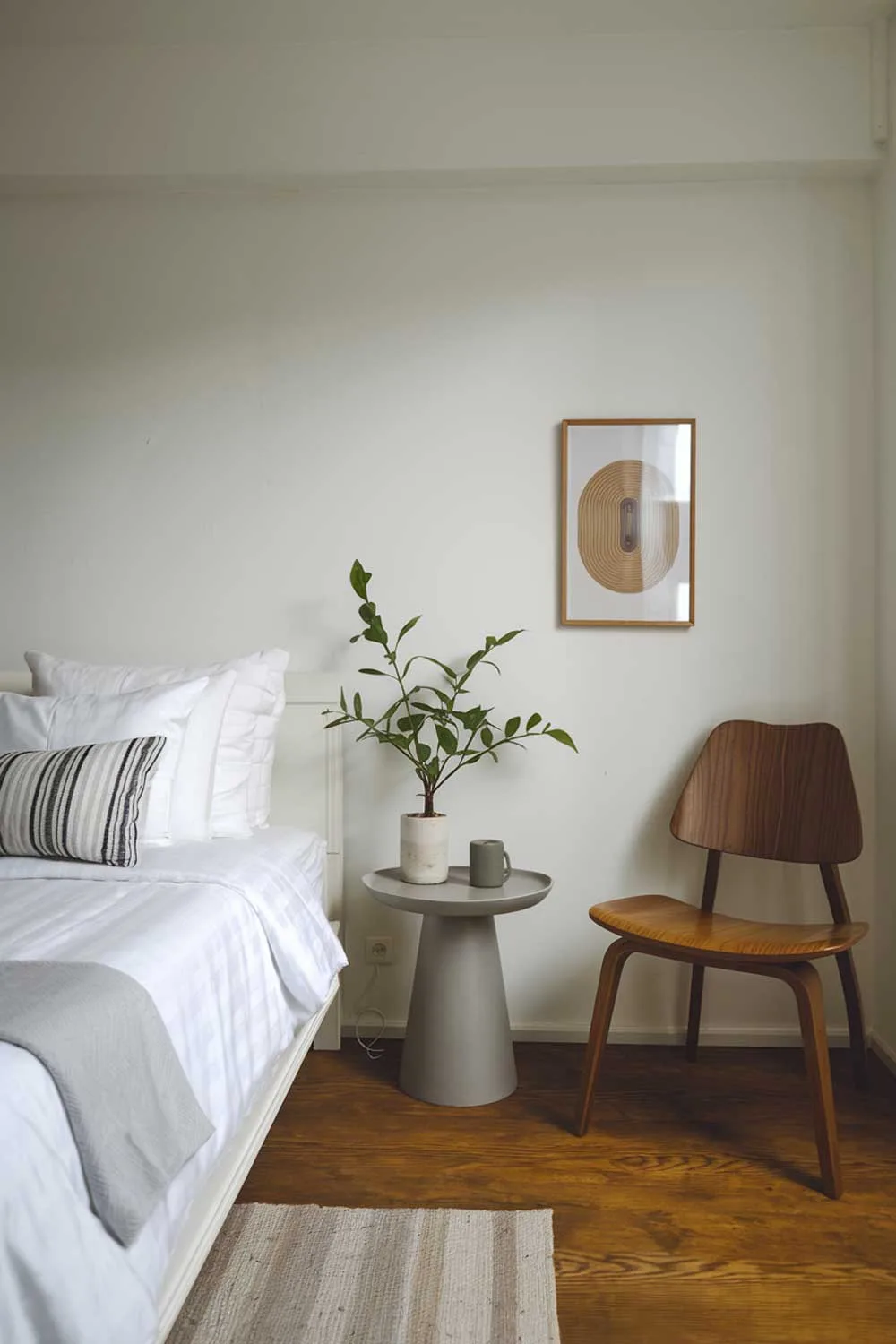 A minimalist apartment bedroom designed for couples. The room is cozy and beautifully designed with a white bed, a gray side table, and a wooden chair. There's a small plant on the side table. The room has a white wall and a wooden floor.