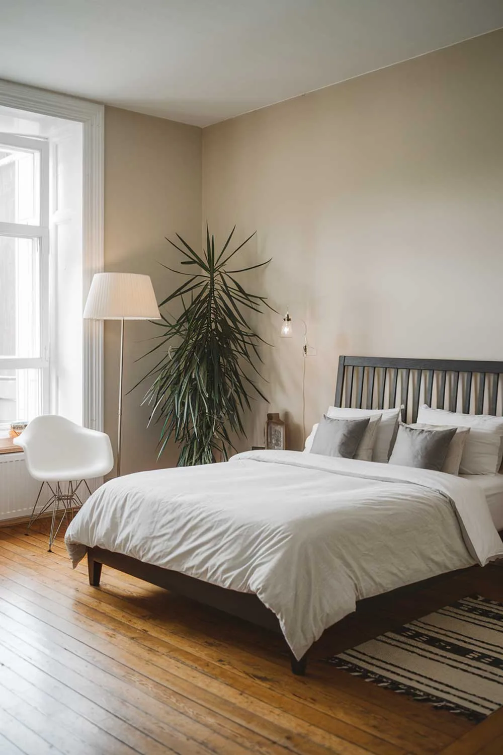 A cozy and beautifully designed minimalist apartment bedroom for couples. The room is spacious and has a king-size bed with a white duvet and gray pillows. A black wooden bed frame stands against a white wall. There's a tall plant in the corner. A floor lamp with a white shade is placed near the bed. A white chair with a black base is positioned under the window. The hardwood floor is visible. The walls are painted beige.