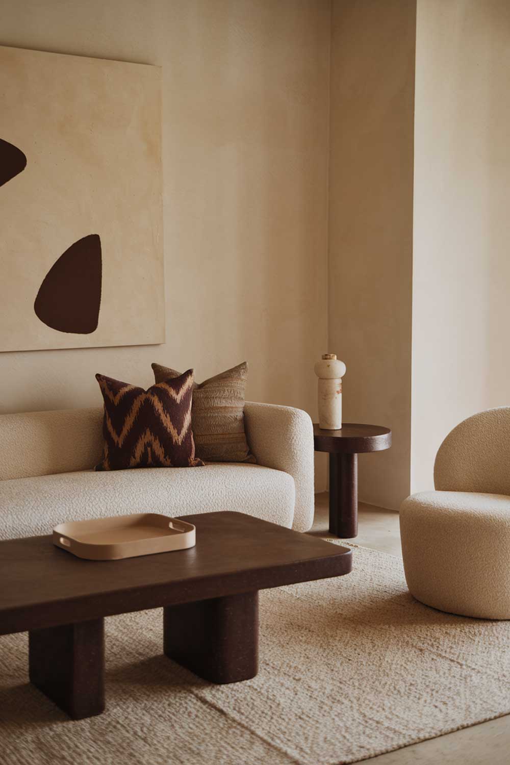  A cozy minimalist living room with earth tones. There is a beige sofa with a few throw pillows in a darker brown and beige pattern. Next to the sofa is a dark brown coffee table with a beige tray on top. A few feet away is a white chair. On the wall is a large beige painting with a few dark brown shapes. The floor is covered with a beige rug.