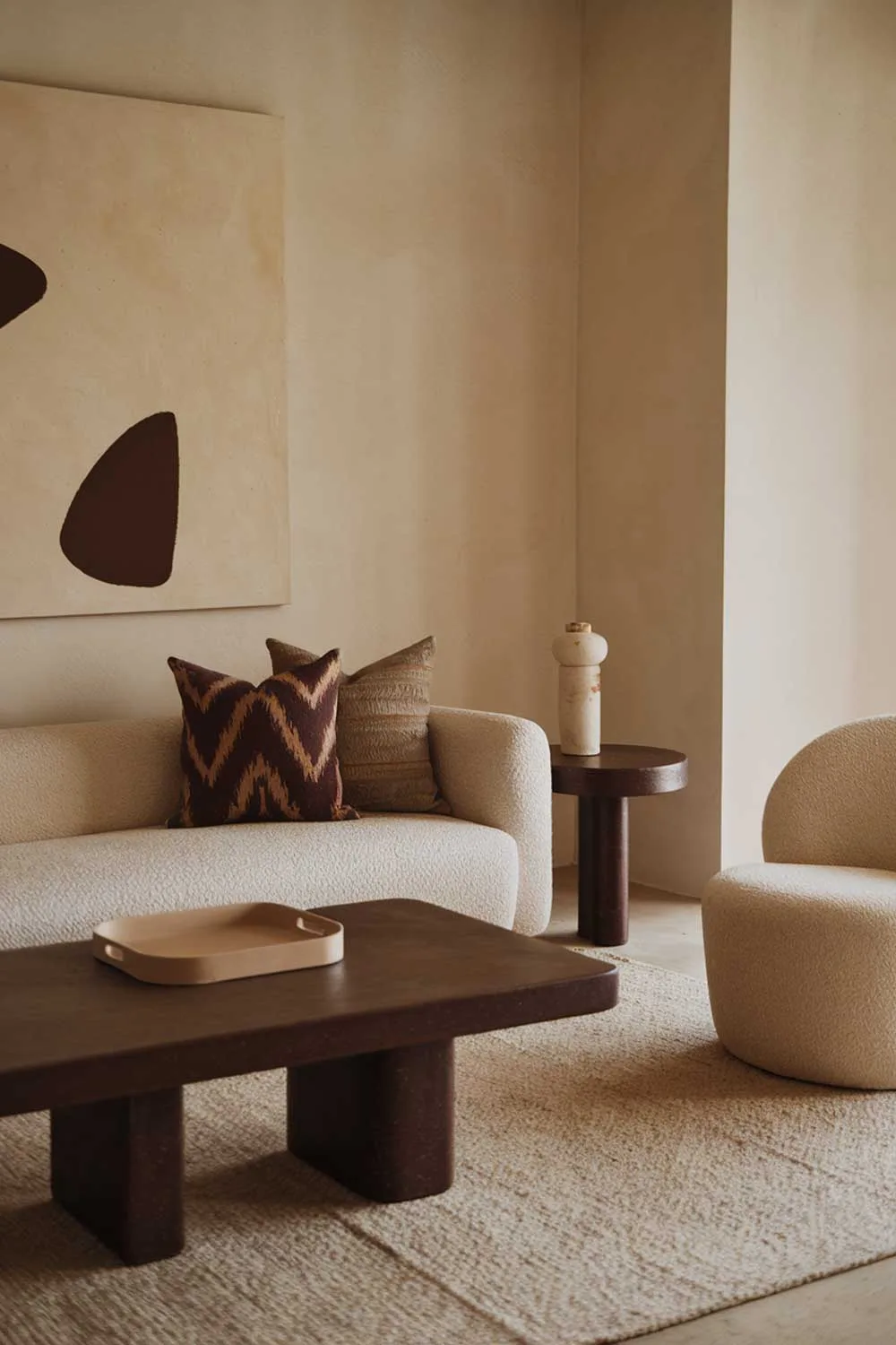 25 Inspiring Minimalist Living Room Ideas with Earth Tones 3 A cozy minimalist living room with earth tones. There is a beige sofa with a few throw pillows in a darker brown and beige pattern. Next to the sofa is a dark brown coffee table with a beige tray on top. A few feet away is a white chair. On the wall is a large beige painting with a few dark brown shapes. The floor is covered with a beige rug.