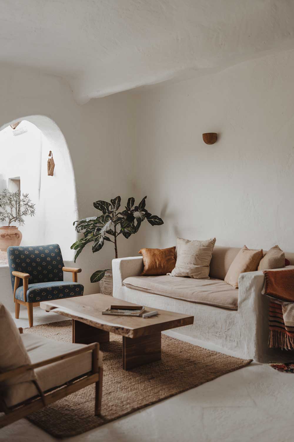 A cozy white minimalist living room with earth tones. There is a beige sofa with a few cushions. There is a blue chair with a pattern. There is a wooden coffee table. There is a potted plant near the window. The walls are white. There is a rug on the floor.