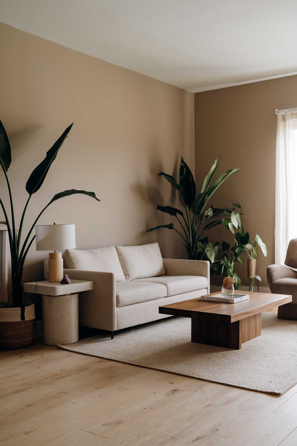 A modern minimalist living room with earthy tones. The room has a beige sofa, a few green plants, and a wooden coffee table. There is a white side table with a lamp next to the sofa. The walls are painted beige. The floor is made of light wooden planks.