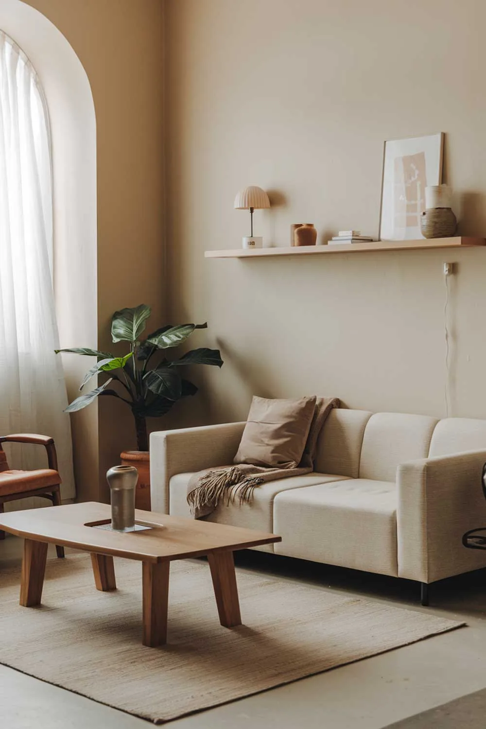 25 Inspiring Minimalist Living Room Ideas with Earth Tones 15 A modern minimalist living room with earth tones. The room has a beige sofa with a few throw pillows. There's a wooden coffee table in the middle of the room. A potted plant is placed near the window. The walls are painted in a light beige color. There's a rug in front of the sofa. A few items are placed on the wall shelf, including a lamp and a few decorative objects.