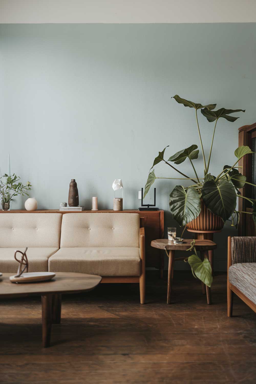 A japandi minimalist living room with earth tones. The room has a beige sofa, a few wooden furniture pieces, and a green plant. The walls are painted in a light blue hue. There is a wooden coffee table and a few decorative items. The floor is made of hardwood.