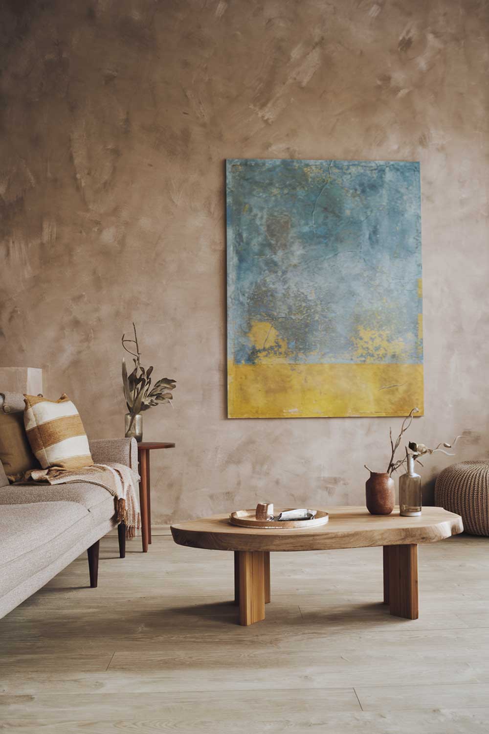 A Scandinavian minimalist living room with earthy tones. The room has a beige sofa with a few throw pillows. There's a wooden coffee table in the middle of the room. On the wall, there's a large, abstract painting with a blue and yellow palette. The floor is made of light wood planks.