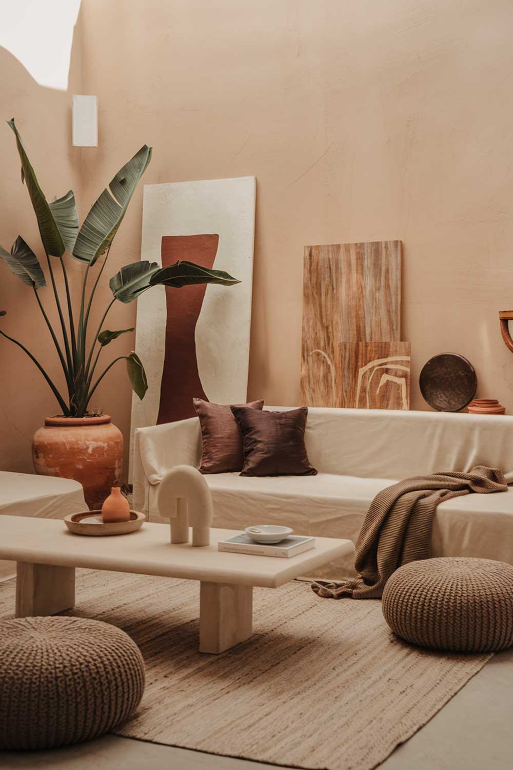 A cozy minimalist living room with earth tones. The room has a beige sofa, a few cushions in darker tones, a white coffee table, and a few decorative items. There's a large plant in a terracotta pot beside the sofa. The floor is covered with a beige rug. The walls have a few pieces of artwork. The lighting is soft and warm.