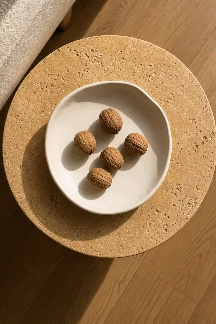 Overhead view of a shallow ivory dish holding five foraged walnuts on a travertine side table in bright midday light