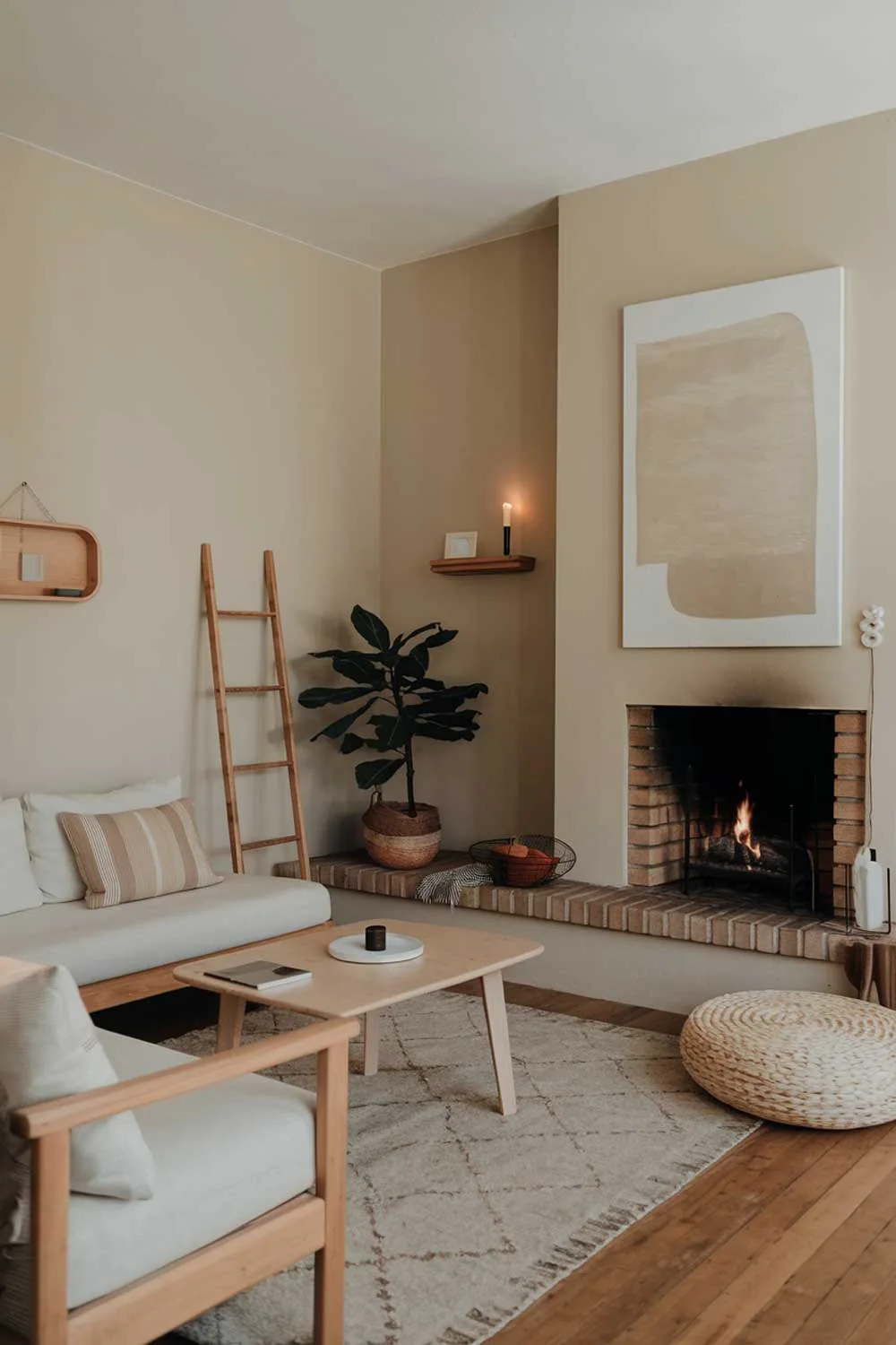 A cozy minimalist living room with a fireplace. There's a white sofa with a few pillows, a wooden coffee table, and a plant. A rug is placed in front of the fireplace. The walls are painted beige, and there are a few pieces of wall art, including a large abstract painting and a small wooden shelf with a candle. There's a wooden ladder next to the wall. The floor is made of wood.