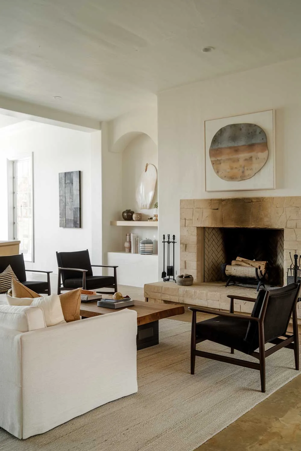 A photo of a cozy minimalist living room with a fireplace. The room has a white sofa and a few black chairs. There's a wooden coffee table in the middle. The walls are adorned with a few pieces of art. The floor is covered with a beige rug. The fireplace is made of stone and has a few logs inside.