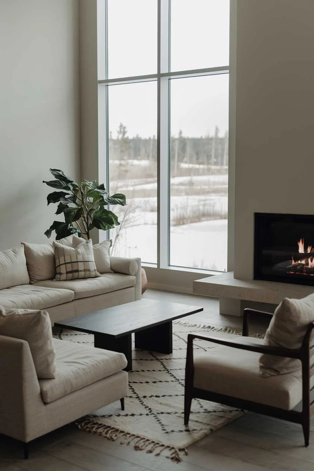 A cozy modern minimalist living room with a fireplace. The room has a large window with a view of a snowy landscape. There's a comfortable beige sofa with cushions and a matching armchair. A dark wooden coffee table is placed in front of the sofa. A rug with a geometric pattern is placed under the furniture. A potted plant is placed near the window. The walls are painted a soft gray color.