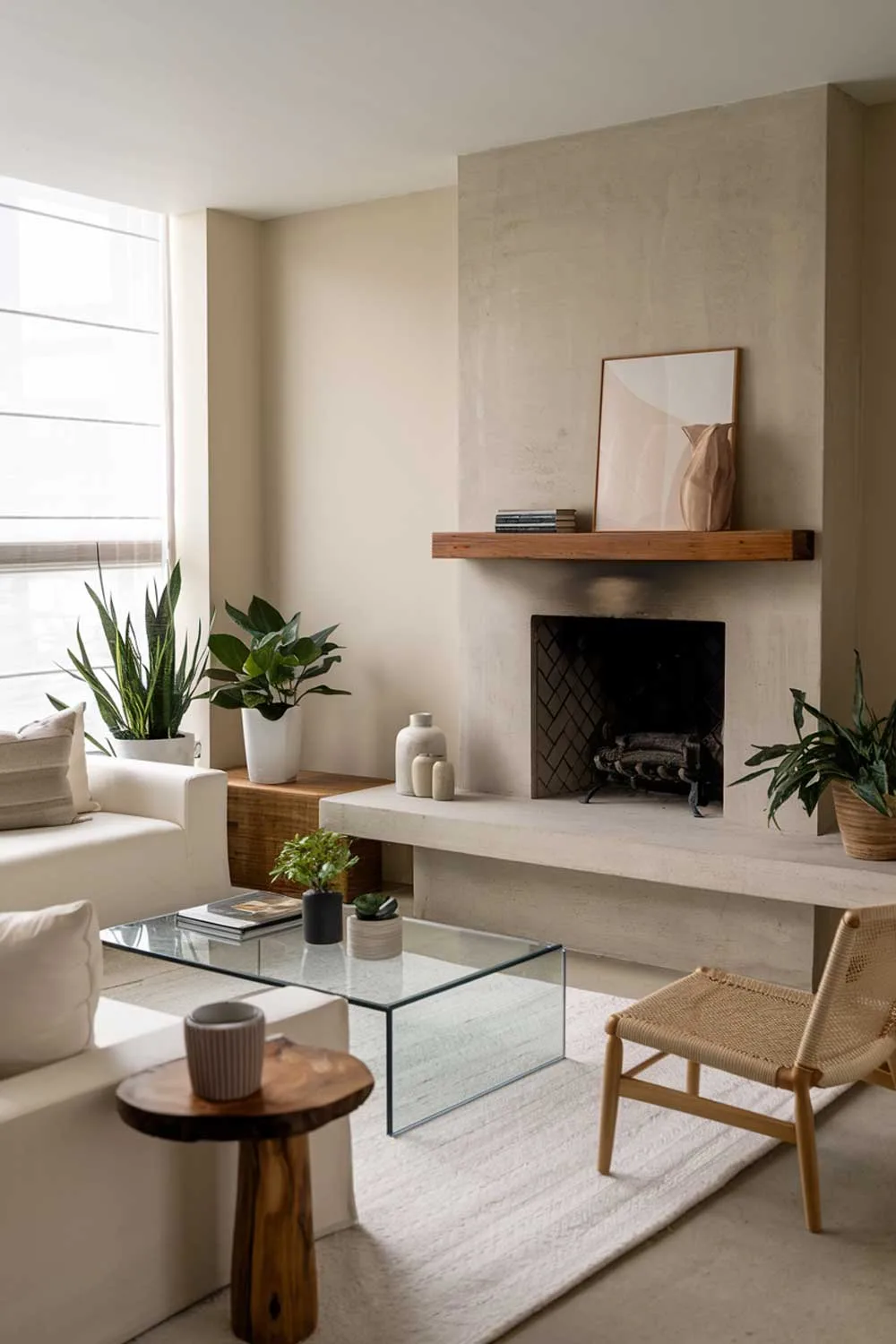 A modern minimalist living room with a cozy fireplace. The room has a white sofa, a few cushions, a glass coffee table, and a wooden side table. A few potted plants are placed near the window. The fireplace is made of concrete and has a wooden shelf above it. The floor is covered with a white rug. The walls are painted beige.