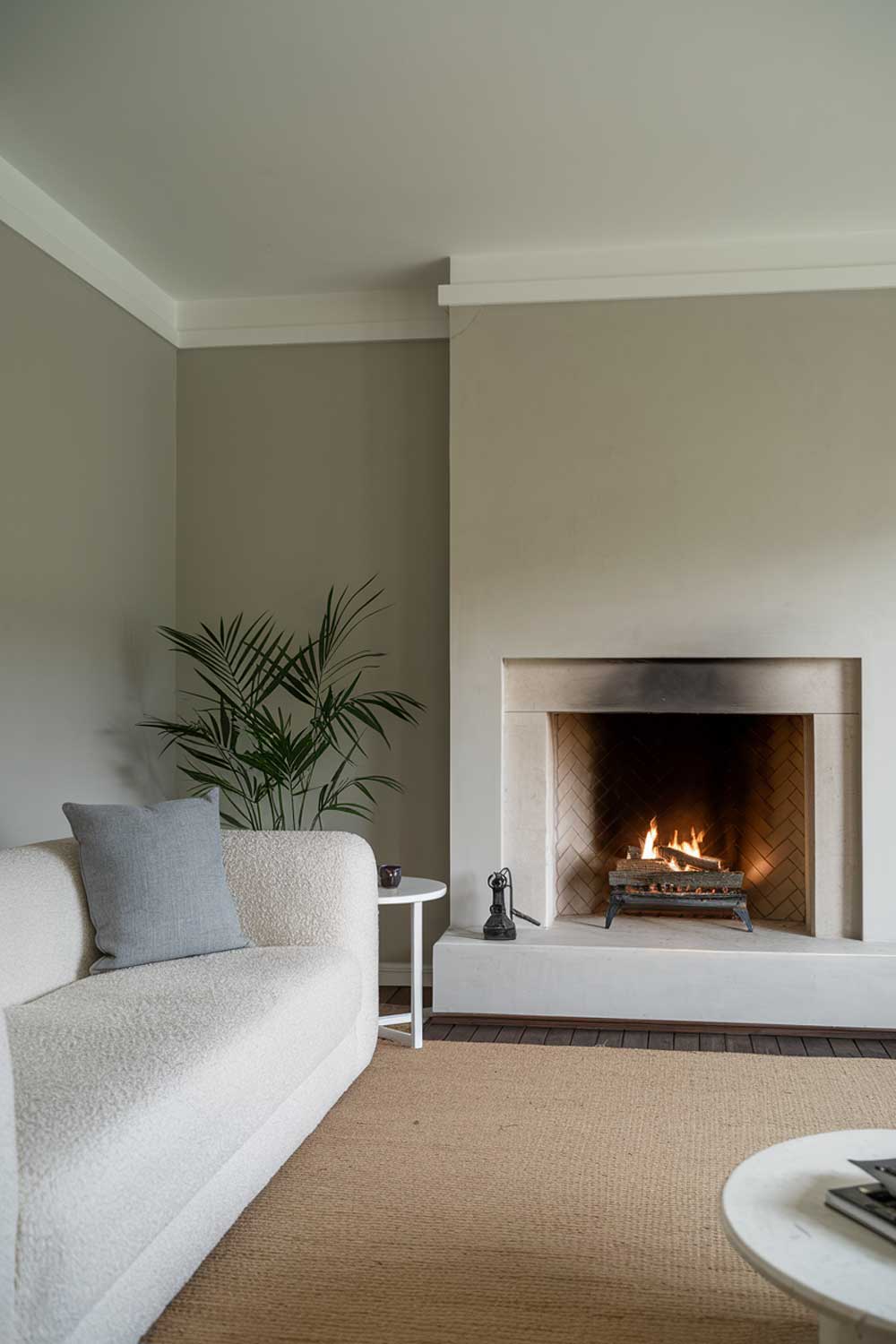 A modern minimalist living room with a cozy fireplace. There's a plush white sofa with a gray throw pillow beside the fireplace. A small white side table sits beside the sofa. A potted plant is placed on the floor. The room has a beige rug. The walls and the fireplace are painted in a light gray color. The ceiling is white.