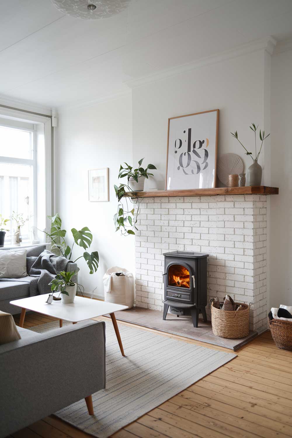 A Scandinavian minimalist living room with a cozy fireplace. The room has a grey sofa, a white coffee table, and a few potted plants. There is a wooden shelf above the fireplace with a few decorative items. The walls and the ceiling are white. The floor is made of wooden planks.