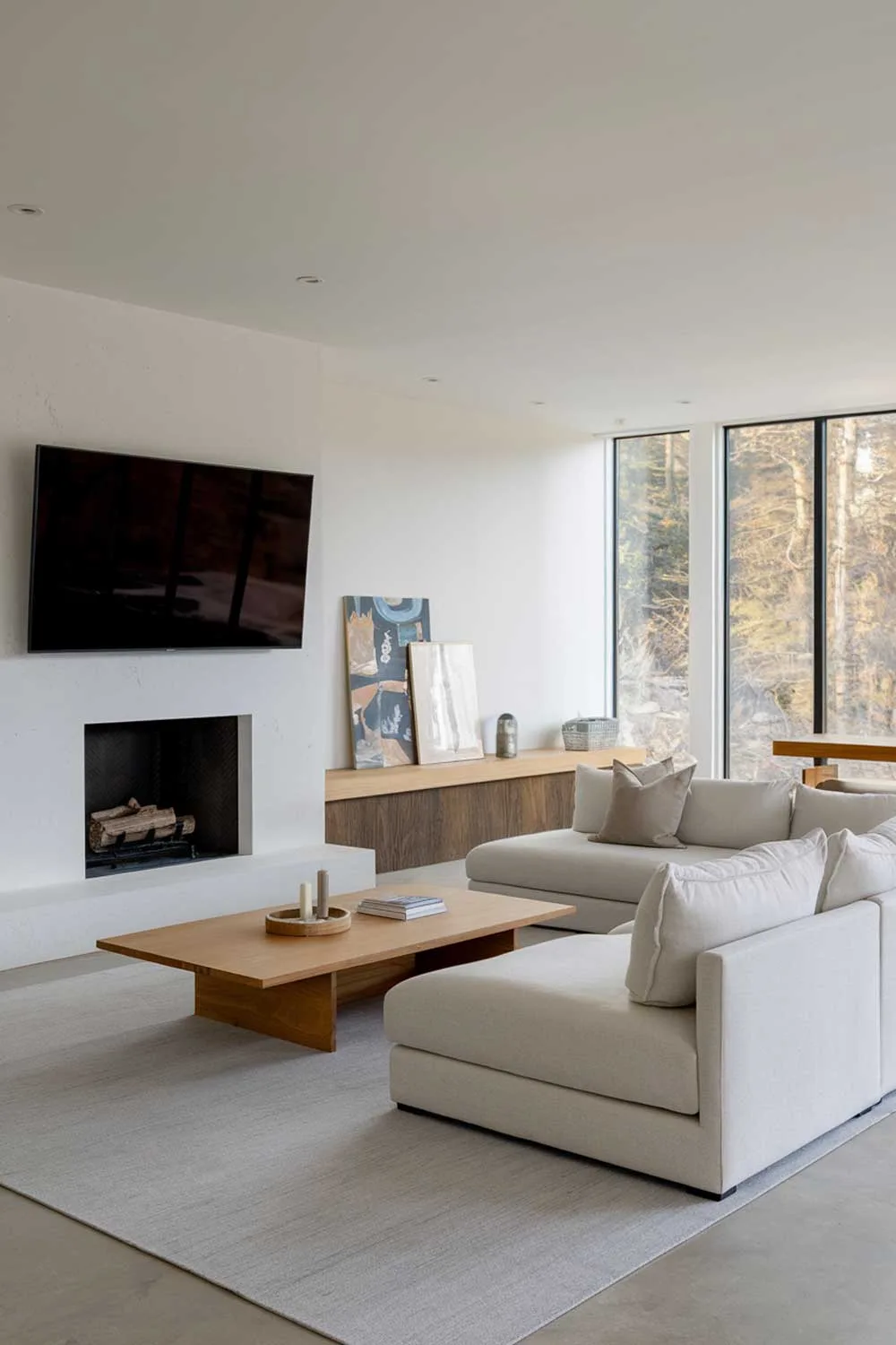 A minimalist living room with a white fireplace and a large TV mounted on the wall. The room has a light grey sectional sofa with a few pillows. There's a wooden coffee table in front of the sofa. The floor is covered with a light grey rug. The wall behind the sofa has a few pieces of artwork. The room has large windows with a view of nature.